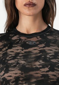 Μαύρη τοπ από δαντέλα με διάφανη, υφασματική υφή, διαθέτοντας στρογγυλή λαιμόκοψη και μια μικρή γκρίζα λογότυπο κοντά στη λαιμόκοψη.