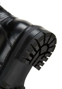 Botte en cuir noir avec une semelle en caoutchouc texturée à crampons profonds, tige lisse contrastante et coutures renforcées le long des coutures.