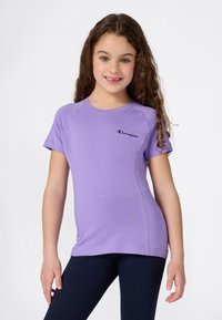 Fille aux longs cheveux bouclés portant un t-shirt violet Champion et un legging noir, souriant devant un fond blanc uni.