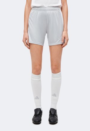 Személy világosszürke Adidas sport rövidnadrágot, fehér, csíkos térdzoknit és fekete focicipőt visel, egyszínű háttér előtt áll.
