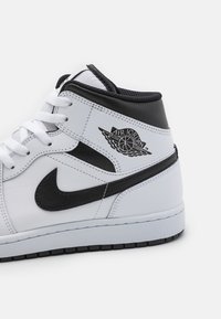 Basket montante Air Jordan blanche et noire montrant une vue latérale avec le logo Nike Swoosh et le logo Air Jordan Wings près de la cheville.