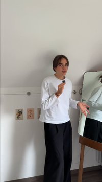 Camisa branca de manga longa, calças pretas de perna larga, em pé à frente de um espelho. A parede exibe imagens com temática botânica. Condições de luz natural.
