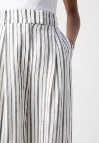 Personne portant un pantalon blanc taille haute avec des rayures verticales noires, des plis, des passants de ceinture, la main dans la poche droite sur un fond uni.