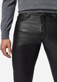 Zwarte leren broeken met een gladde afwerking, gedetailleerde stiksels en metalen hardware. Gestyled met een aangesloten ontwerp. Grijze trui zichtbaar.