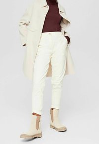 Cappotto beige sopra un dolcevita bordeaux, abbinato a pantaloni color crema e stivaletti beige con pannelli laterali. Design minimalista.