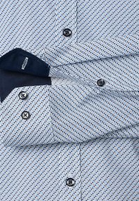 Koszula na guziki w jasnym niebieskim kolorze z geometrycznym wzorem. Zawiera ciemne guziki i granatowy wewnętrzny kołnierzyk. Wykonana z bawełny, o gładkiej teksturze.