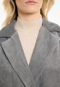 Grijze corduroy jas met brede revers, gecombineerd met een beige geribde coltrui. Heeft verticale ribbels en een zachte textuur.