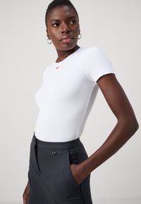 White ribbed t-shirt with short sleeves and a small red logo at the neck, paired with dark tailored trousers featuring a side pocket and button.