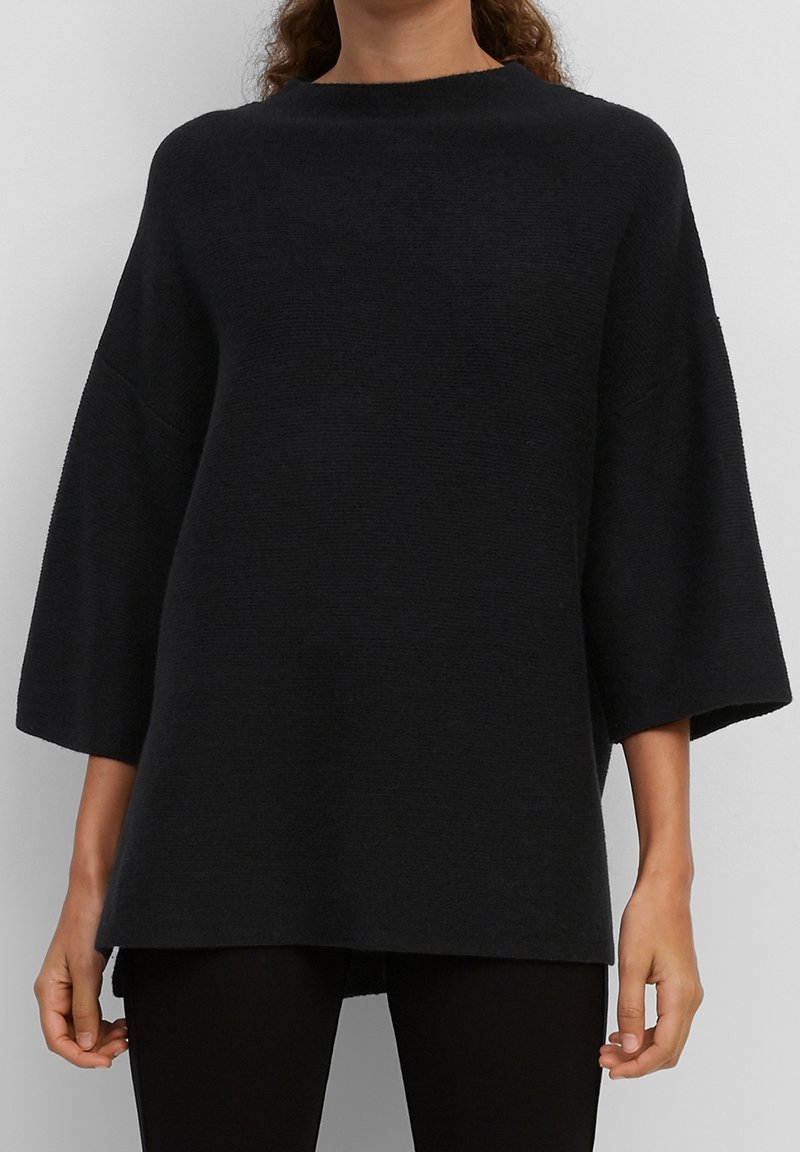 Femme portant un pull en maille noire ample avec de larges manches trois-quarts et un col rond haut, associé à un pantalon noir.