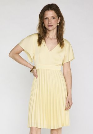 Femme aux cheveux bruns ondulés portant une robe plissée jaune clair à manches courtes, des boucles d'oreilles créoles dorées et des bracelets en or, debout devant un fond uni.