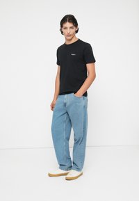Tricou negru din bumbac, cu mânecă scurtă și un logo mic, purtat cu blugi din denim de culoare albastru deschis și adidași albi cu tălpi bej.