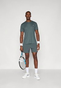 T-shirt desportivo verde-escuro e calções combinando, com braceletes brancas e raquete de ténis, apresentando um detalhe azul no cabo da raquete.