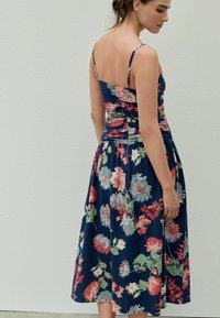 Blumenkleid mit Spaghettiträgern, auf marineblauem Hintergrund, mit leuchtend roten, rosa und grünen Blumen. Raffung am Oberteil und fließendes Rockdesign.