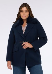 Navyblauer Mantel mit gefälschtem Pelzkragen, großen Revers und tailliertem Gürtel. Verfügt über zwei Vordertaschen und einen weichen, strukturierten Stoff.