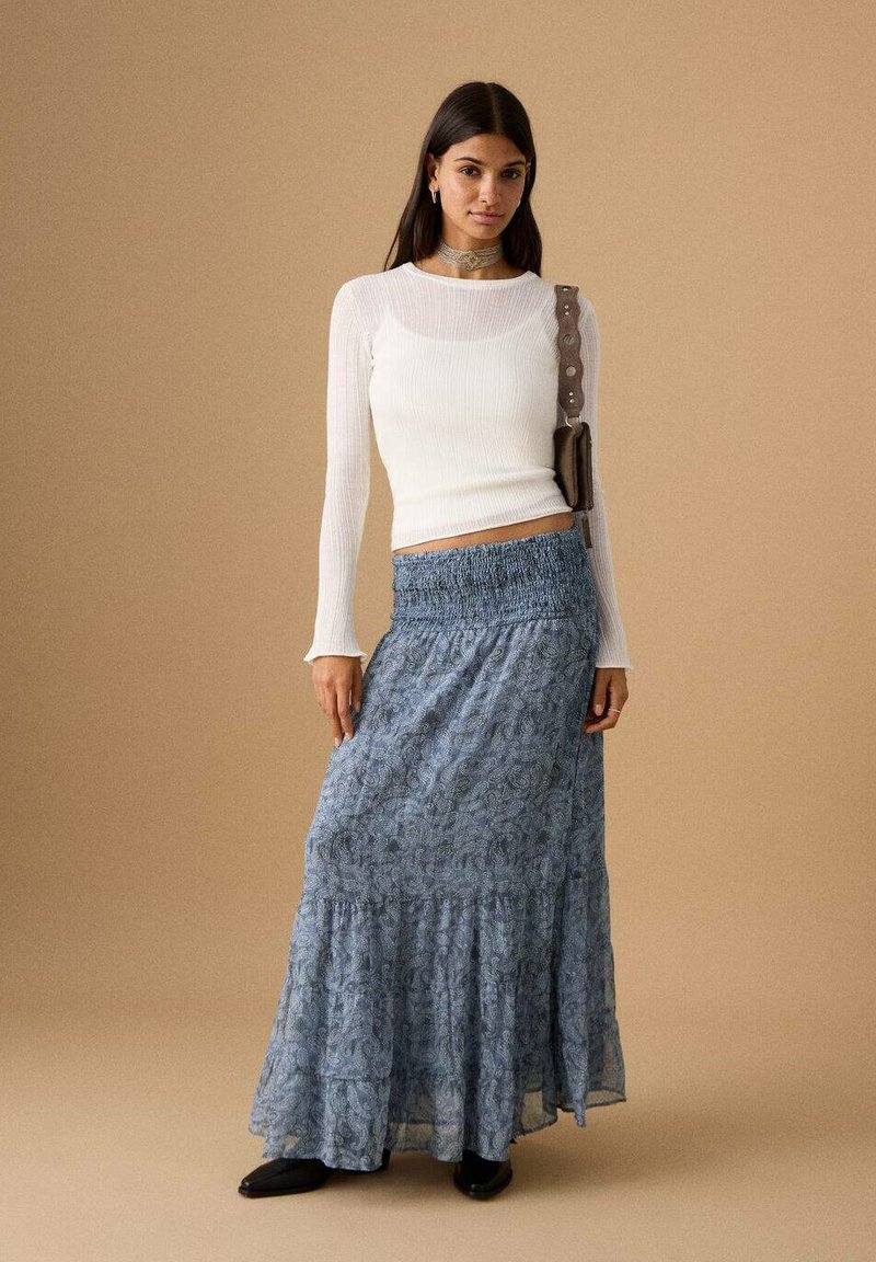 Top bianco a maniche lunghe a coste abbinato a una gonna lunga blu con fantasia. La gonna presenta una vita arricciata e un orlo a balze. Scarpe nere.