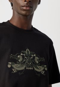 Nærbilde av en svart skjorte med grønne broderte fugler, blomster og blader, samt ordet "HUGO" sentrert under designet.