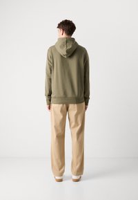 Felpa verde oliva con cappuccio regolabile, polsini e orlo a coste. Abbinata a pantaloni beige a gamba larga e scarpe bianche.