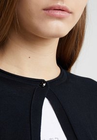 Černý pletený kardigan s kulatým výstřihem, který má jedno černé zapínání na knoflík a texturovanou tkaninu. Pod ním je vidět bílá košile.