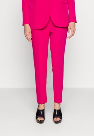 Personne portant un pantalon rose vif sur mesure et un blazer assorti, associé à des chaussures noires à talons ouverts, debout sur un sol simple.