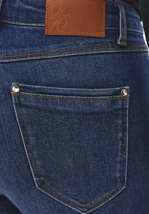 Primer plano del bolsillo trasero de unos vaqueros de mezclilla azul oscuro con remaches dorados y una etiqueta de marca de cuero marrón cosida encima del bolsillo.