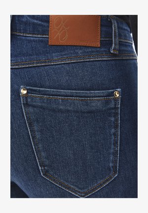 Primer plano del bolsillo trasero de unos vaqueros de mezclilla azul oscuro con remaches dorados y una etiqueta de marca de cuero marrón cosida encima del bolsillo.