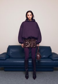 Felpa oversize viola, minigonna con paillettes, leggings viola, stivali alti con punta, e una piccola borsa marrone, su un divano blu.