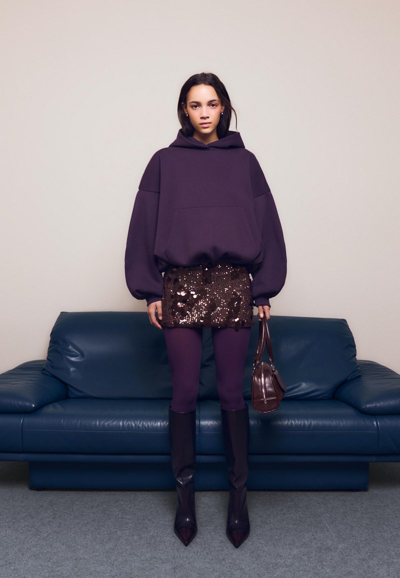 Felpa oversize viola, minigonna con paillettes, leggings viola, stivali alti con punta, e una piccola borsa marrone, su un divano blu.