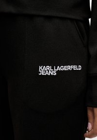 Primo piano di una tasca di pantaloni neri con il logo bianco ricamato "KARL LAGERFELD JEANS" e una parziale visione di una mano appoggiata accanto alla tasca.