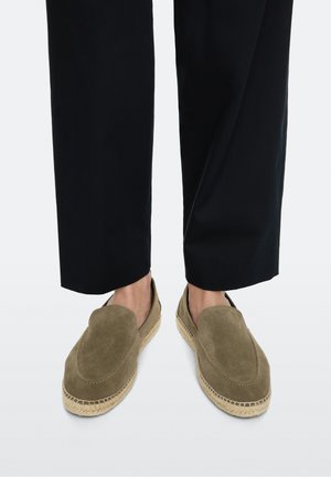 Person, die olivgrüne Wildleder-Slip-on-Espadrilles mit gewebten Jutesohlen und schwarze verkürzte Hosen auf hellem Hintergrund trägt.