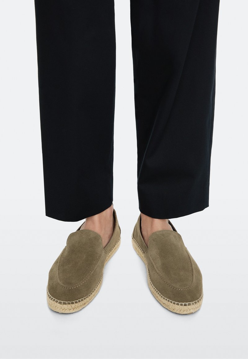 Person, die olivgrüne Wildleder-Slip-on-Espadrilles mit gewebten Jutesohlen und schwarze verkürzte Hosen auf hellem Hintergrund trägt.