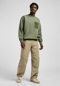 Groene sweatshirt met een zak, gecombineerd met beige broek met brede pijpen en lichtgekleurde sneakers. De stof lijkt zacht en heeft een textuurafwerking.