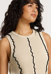 Beidzis adīts bezpiedurkņu topiņš ar teksturētu rakstu. Priekšpusē ir melni viļņaini akcenti. Apaļa kakla izgriezums.