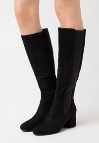 Bottes montantes en daim noir avec talons carrés portées sur des jambes nues, dotées d'une fermeture éclair latérale et d'un panneau élastique pour ajuster la coupe.