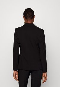Blazer nero con bavero strutturato, design aderente e maniche lunghe. Tessuto liscio con retro sartoriale, abbinato a pantaloni neri.