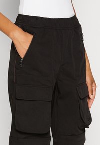 Černé nákladní kalhoty z odolné látky, s elastickým pasem, bočním zipovým kapsou a dvěma velkými kapsami s klopami na stehnech.