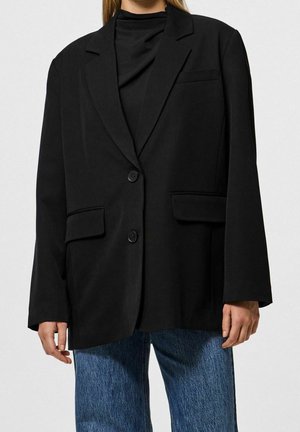 Kobieta ubrana w za duży czarny blezer z dwoma guzikami i przednimi kieszeniami z patką, na czarnym topie i niebieskich dżinsach.