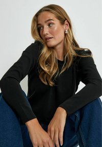 Top nero a maniche lunghe realizzato in morbido tessuto, con scollatura rotonda. Il modello indossa jeans in denim blu, con le mani appoggiate sulle ginocchia e capelli mossi e naturali.