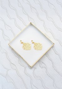 Guldtonade metallörhängen med intrikat blommönster, visade i en fyrkantig vit låda med guldkanter på en texturerad yta.