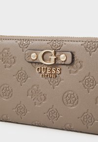 Beige textuur portemonnee met ingebracht GUESS-logo, gouden hardware en een ritsluiting. Kenmerkt een elegante geometrische patroon en een gladde afwerking.