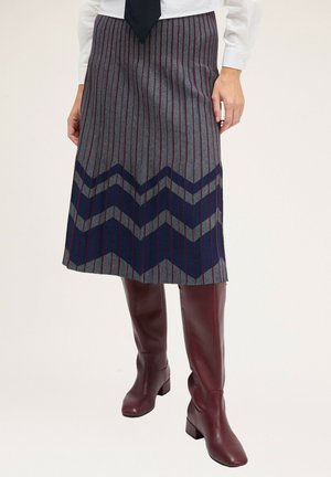 Gonna di lana grigia con strisce rosse verticali e motivo a zigzag blu navy all'orlo, indossata con stivali in pelle bordeaux alti fino al ginocchio.