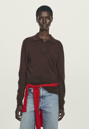 Woman wearing a dark brown long-sleeve polo sweater with a red sweater tied around her waist and light blue jeans against a plain background.