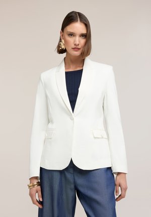 Donna che indossa un blazer bianco monopetto su una maglia a coste scura e jeans blu larghi, accessoriata con orecchini e braccialetti dorati.