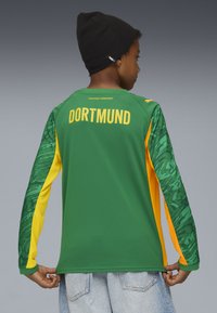 Lapsi, joka pitää yllään vihreän ja keltaisen Dortmund-paidan sekä mustan Puma-pipon, kääntää selkänsä ja pitää paidan helmaa kiinni, harmaata taustaa vasten.