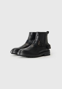 Botas de cuero negro hasta el tobillo con punteras redondas, suelas gruesas y correas laterales con hebillas de metal, presentadas sobre un fondo blanco.