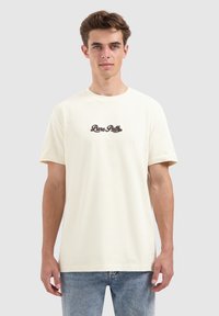 Jonge man met kort bruin haar, gekleed in een crèmekleurig T-shirt met de tekst "Pure Patti" en lichtblauwe jeans, staande tegen een effen achtergrond.