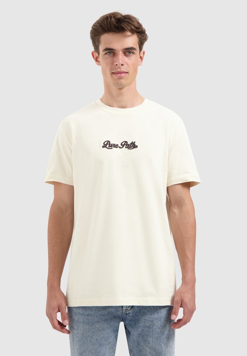 Jonge man met kort bruin haar, gekleed in een crèmekleurig T-shirt met de tekst "Pure Patti" en lichtblauwe jeans, staande tegen een effen achtergrond.