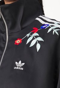 Svart träningsjacka med blommig brodyr i röd, blå och grön. Inkluderar en silverdragkedja och tre vita ränder på axeln.