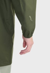 Manche de veste vert olive avec poignet élastique ; comporte un logo subtil imprimé dans une teinte plus claire sur le tissu. Main posée à proximité.