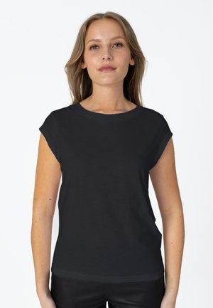 Femme aux cheveux châtain clair portant un t-shirt noir uni à manches courtes, debout devant un fond blanc.