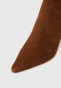 Bota de tobillo de ante marrón con punta afilada, textura suave y silueta estilizada. Cuenta con un tacón de aguja y un diseño minimalista.